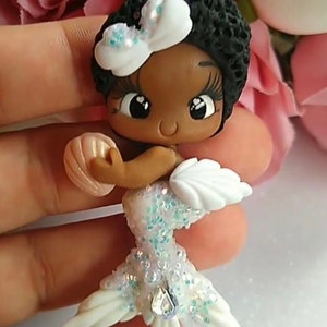 May include: A handcrafted mermaid figurine with dark skin, black curly hair, and a white bow. The mermaid's tail is white with iridescent glitter and a central jewel. She holds a seashell and has white fins.