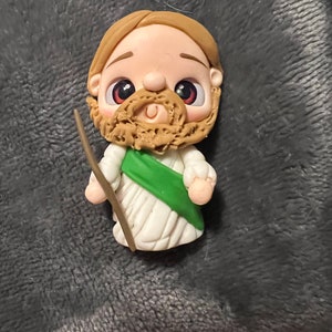 May include: A handcrafted figurine of a bearded man with a staff. The figure has a light skin tone, brown hair, and a tan beard. He wears a white robe with a green sash. The figurine is made of clay.