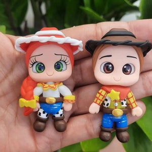 May include: Two miniature figurines of cartoon characters. One is a cowgirl with a red hat, red hair, and blue pants. The other is a cowboy with a brown hat, yellow shirt, and blue pants. Both have large eyes and are smiling.