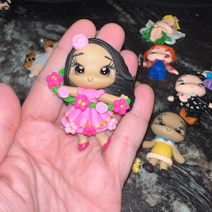 May include: A handcrafted figurine of a cartoon character with dark hair, wearing a pink dress and flower crown. The figurine is held in a hand, with other small figurines in the background.