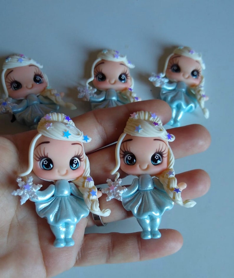 K&ouml;nnte beinhalten: Eine Sammlung von f&uuml;nf Miniaturfiguren. Jede Figur hat ein hellblaues Kleid, blondes Haar und eine sternf&ouml;rmige Krone. Die Figuren halten schneeflockenf&ouml;rmige Objekte. Die Gesichter haben gro&szlig;e, ausdrucksstarke Augen.