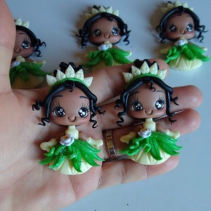 May include: Handmade figurines of a princess with dark skin, black curly hair, and a white and green crown. The princess wears a green and yellow dress with a white flower accent. The figurines are held in a hand.