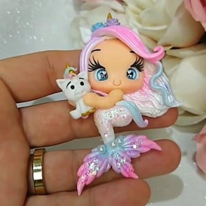 May include: A handcrafted mermaid figurine with a cat. The mermaid has pink and blue hair, blue eyes, and a glittery tail in shades of pink, blue, and purple. The cat is white with a golden horn. The figurine is held in a hand.