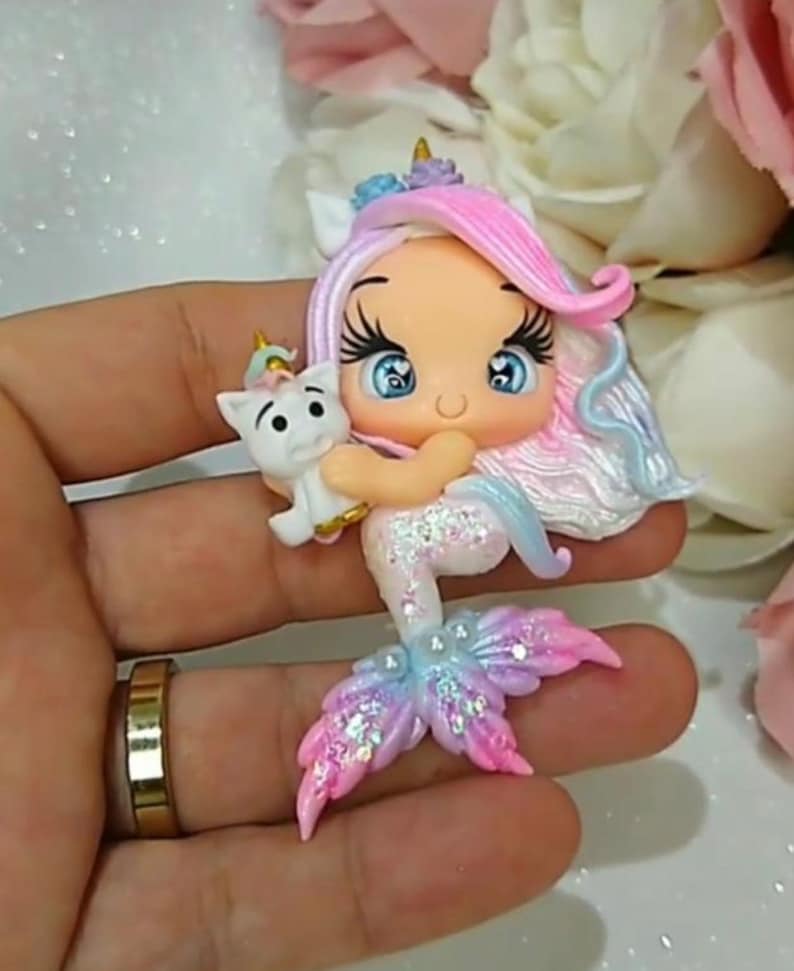 May include: A handcrafted mermaid figurine with a cat. The mermaid has pink and blue hair, blue eyes, and a glittery tail in shades of pink, blue, and purple. The cat is white with a golden horn. The figurine is held in a hand.