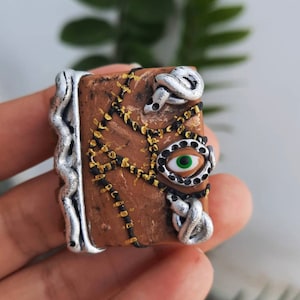 May include: A miniature brown book with a silver spine and accents. The book features a green-eyed design and silver snake-like embellishments. Gold stitching and black lines add detail to the cover.