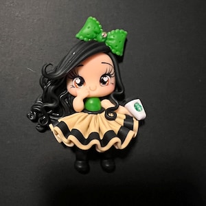 May include: A handcrafted figurine of a girl with black hair, a green bow, and a tan and black dress. The figurine is holding a small white cup with a green logo. The girl has large eyes and is smiling.