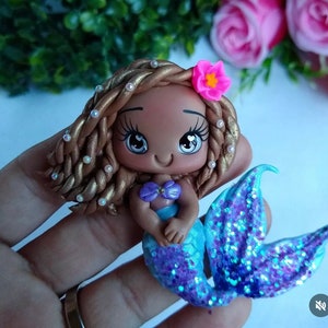 May include: A handcrafted mermaid figurine with a brown complexion, adorned with gold-colored hair and a pink flower. The mermaid's tail is a shimmering blue with glitter, and she wears a purple seashell top. The figurine is a charming decorative item.