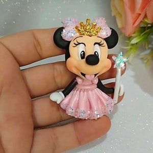 May include: A Minnie Mouse figurine wearing a pink dress with glitter accents and a gold crown. The figure holds a star-shaped wand. The character's black ears and bow are adorned with pink glitter.