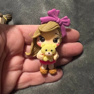 May include: A handmade polymer clay figurine of a girl with light brown hair, a purple bow, and a yellow teddy bear. The girl wears a pink skirt and brown shoes. The figurine is held in a hand against a gray background.