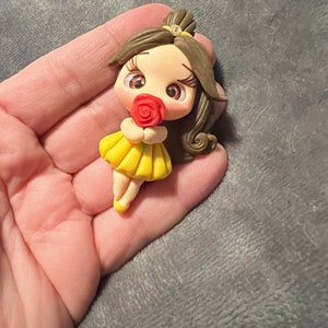 May include: A miniature figurine of a cartoon character with brown hair, a yellow dress, and a red rose. The figurine has large eyes and is holding the rose. The character is a princess.