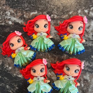 Puede incluir: Cinco figuras hechas a mano de un personaje de dibujos animados con cabello rojo, un vestido verde y un p&aacute;jaro amarillo. Cada figura tiene una flor rosa en el pelo y una falda brillante azul y verde. Las figuras est&aacute;n sobre una superficie gris oscuro.