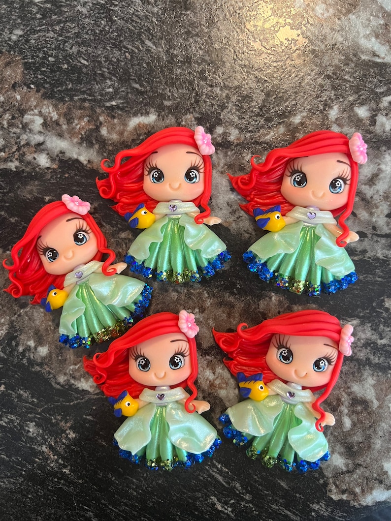 Puede incluir: Cinco figuras hechas a mano de un personaje de dibujos animados con cabello rojo, un vestido verde y un p&aacute;jaro amarillo. Cada figura tiene una flor rosa en el pelo y una falda brillante azul y verde. Las figuras est&aacute;n sobre una superficie gris oscuro.