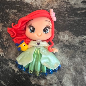 Puede incluir: Figura de Ariel con cabello rojo, una flor rosa y un vestido verde. La figura tiene ojos grandes y una expresi&oacute;n sonriente. Un pez amarillo y azul est&aacute; unido a la figura. El vestido tiene purpurina azul en el dobladillo.