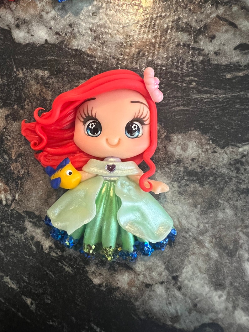 Puede incluir: Figura de Ariel con cabello rojo, una flor rosa y un vestido verde. La figura tiene ojos grandes y una expresi&oacute;n sonriente. Un pez amarillo y azul est&aacute; unido a la figura. El vestido tiene purpurina azul en el dobladillo.
