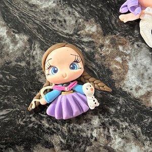 May include: A handcrafted polymer clay figurine of a character with blue eyes, brown braids, and a purple skirt, holding a small snowman. The figurine has a blue top and a pink scarf. The overall design is whimsical and detailed.