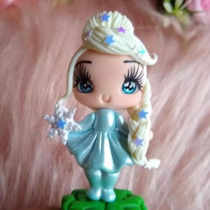 K&ouml;nnte beinhalten: Figur einer Zeichentrickfigur mit hellblauem Kleid und Haar. Die Figur hat lange Wimpern, blaue Augen und ein kleines L&auml;cheln. Das Haar ist zu einem Zopf und Dutt frisiert, verziert mit blauen sternf&ouml;rmigen Dekorationen. Die Figur h&auml;lt eine Schneeflocke und steht auf einem gr&uuml;nen Sockel.