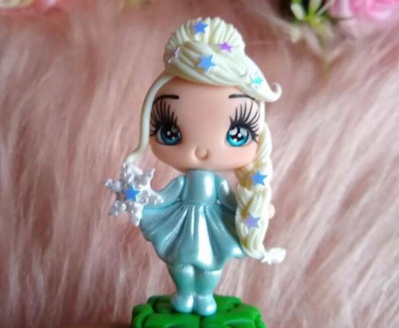 K&ouml;nnte beinhalten: Figur einer Zeichentrickfigur mit hellblauem Kleid und Haar. Die Figur hat lange Wimpern, blaue Augen und ein kleines L&auml;cheln. Das Haar ist zu einem Zopf und Dutt frisiert, verziert mit blauen sternf&ouml;rmigen Dekorationen. Die Figur h&auml;lt eine Schneeflocke und steht auf einem gr&uuml;nen Sockel.