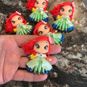 Puede incluir: Colecci&oacute;n de figuras hechas a mano que representan a un personaje de dibujos animados con pelo rojo, un vestido verde y un pez amarillo. Las figuras tienen detalles de purpurina y se exhiben sobre una superficie oscura. Cada figura tiene una expresi&oacute;n &uacute;nica.
