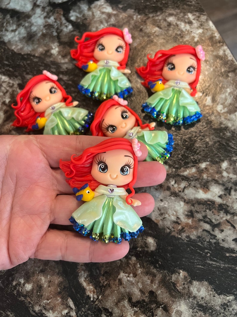 Puede incluir: Colecci&oacute;n de figuras hechas a mano que representan a un personaje de dibujos animados con pelo rojo, un vestido verde y un pez amarillo. Las figuras tienen detalles de purpurina y se exhiben sobre una superficie oscura. Cada figura tiene una expresi&oacute;n &uacute;nica.