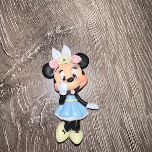 May include: A figurine of Minnie Mouse, featuring black ears, a light blue dress, and yellow shoes. Minnie is adorned with a floral headpiece with pink and white accents. The figurine has a playful expression.