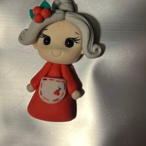 May include: A handcrafted figurine of a woman in a red dress and white apron, adorned with a sprig of holly. The figure has gray hair, black eyes, and a smiling expression. A charming Christmas decoration.