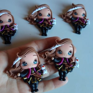 May include: A collection of handmade figurines featuring a cartoon character with brown hair, blue eyes, and a white headpiece. The character wears a black and gold outfit with a purple accent and holds a small snowman. The figurines are displayed on a light surface.