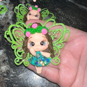 May include: A collection of fairy figurines. Each figurine features a doll-like face with brown hair, a floral crown, and a teal dress. The fairies have glittery green wings. The figurines are made of a polymer clay material.