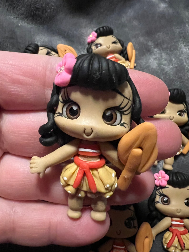 Pode incluir: Uma cole&ccedil;&atilde;o de pequenas figuras artesanais. Cada figura apresenta uma personagem de desenho animado com cabelo escuro, uma flor rosa e uma saia amarela com bolinhas brancas. A personagem segura um objeto castanho.