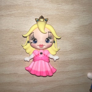 May include: A Princess Peach figurine with a golden crown, blonde hair, and a pink dress. The figurine has large blue eyes and is in a standing pose. The dress has a darker pink trim at the bottom.