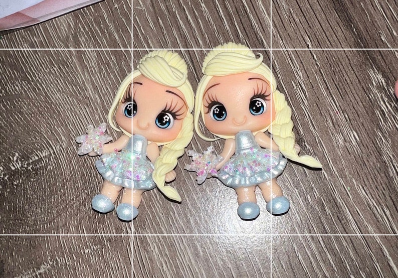 May include: Two miniature doll figures with blonde hair styled in braids. They wear light blue dresses with glitter accents and hold star-shaped accessories. The dolls have large eyes and are posed on a wooden surface.