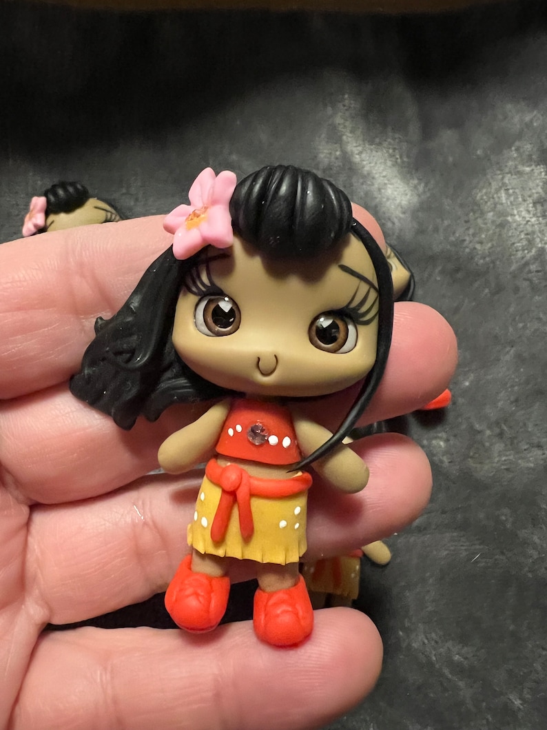May include: A small, handcrafted doll with dark hair and a pink flower. The doll wears a red top with white dots, a yellow skirt with white dots, and orange shoes. The doll's eyes are large and brown.
