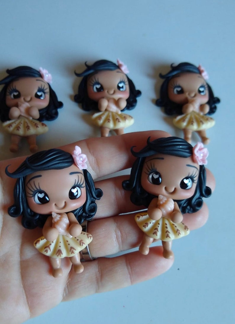 Puede incluir: Figuras hechas a mano de ni&ntilde;as sonrientes con cabello oscuro, piel clara y accesorios para el cabello de flores. Las ni&ntilde;as visten vestidos amarillos con detalles marrones. Las figuras se sostienen en una mano.
