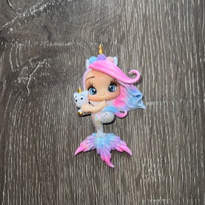 May include: A whimsical figurine of a mermaid with a unicorn horn and flowing pink and blue hair. The mermaid is holding a small white unicorn. The tail is a gradient of pink, blue, and purple with glitter accents.