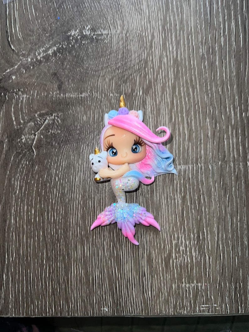 May include: A whimsical figurine of a mermaid with a unicorn horn and flowing pink and blue hair. The mermaid is holding a small white unicorn. The tail is a gradient of pink, blue, and purple with glitter accents.