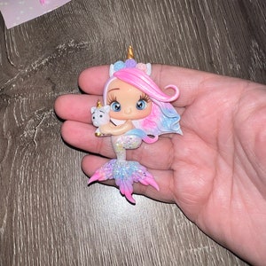 May include: A whimsical figurine of a mermaid-unicorn hybrid, held in a hand. The figure has a pink and blue mane, a golden horn, and a glittery tail. It holds a small white unicorn. The figurine is approximately 3 inches tall.