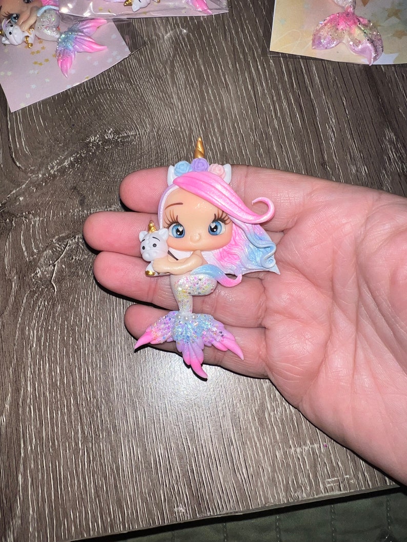 May include: A whimsical figurine of a mermaid-unicorn hybrid, held in a hand. The figure has a pink and blue mane, a golden horn, and a glittery tail. It holds a small white unicorn. The figurine is approximately 3 inches tall.