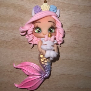 May include: A whimsical figurine of a mermaid-unicorn hybrid, featuring a pink tail and hair, and a golden horn. The mermaid holds a white creature. The figurine is adorned with pastel-colored flowers and has large, expressive eyes.