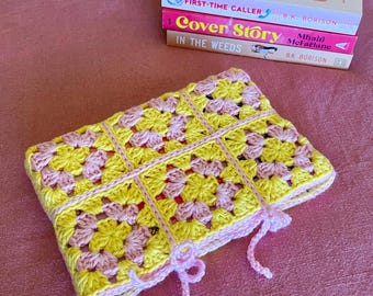 May include: A handmade crochet case in yellow and pink, with a grid pattern and pink ties. The case is next to a stack of three books with the titles "First-Time Caller" and "Cover Story". The books are on a pink surface.