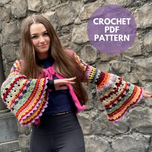 May include: A colorful crochet top with bell sleeves. The top is made with a variety of bright colors, including pink, red, orange, yellow, green, blue, and purple. The sleeves are long and flared, and they have a scalloped edge. The top is tied at the neck with a pink ribbon.
