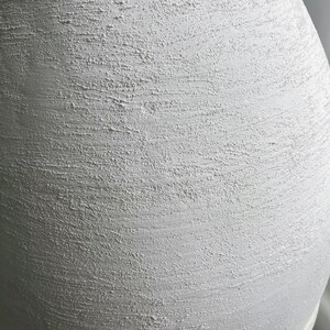 Pode incluir: Um vaso de cer&acirc;mica branco com uma superf&iacute;cie texturizada. O vaso tem uma forma arredondada e uma abertura larga.