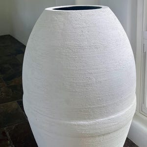 Pode incluir: Um vaso grande de cer&acirc;mica branca texturizada com formato arredondado. O vaso tem uma borda preta lisa.