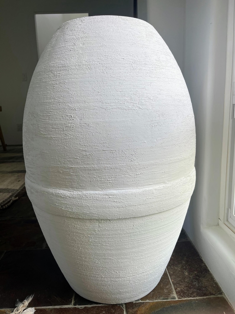 Pode incluir: Um vaso grande, branco, de forma oval e com superf&iacute;cie texturizada. O vaso tem uma faixa ligeiramente elevada ao redor do meio.