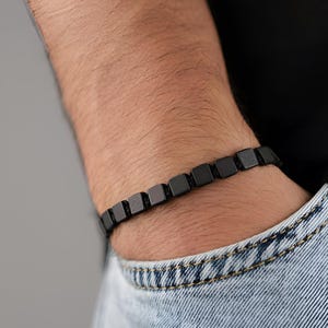 Op de afbeelding: Zwart vierkant kralenarmband. De armband is gemaakt van kleine, gelijkmatig verdeelde, matzwarte kralen die aan elkaar zijn geregen. De armband wordt om een pols gedragen, met een spijkerbroekzak op de achtergrond.