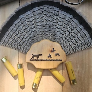 May include: A wooden plaque with a silhouette of a dog pointing at three flying birds. The plaque is surrounded by a turkey tail fan and shotgun shells.