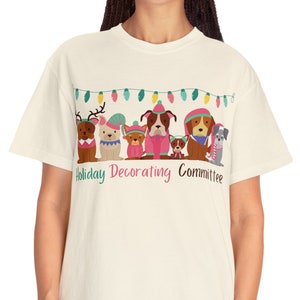 Op de afbeelding: Een crèmekleurig T-shirt met een afbeelding van honden die wintermutsen en sjaals dragen. De tekst "Holiday Decorating Committee" staat onder de afbeelding.
