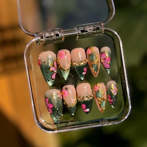 Puede incluir: Un juego de diez uñas postizas en un estuche de plástico transparente, con una mezcla de bases color nude y verde oscuro. Las uñas están decoradas con flores rosas pintadas a mano, detalles metálicos dorados y pedrería incrustada en punta.