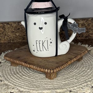May include: A white ceramic Halloween-themed mug with a witch hat and the word "EEK!" in black lettering. The mug has a handle and is decorated with a smiling face. It sits on a small, square wooden tray.