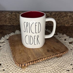 May include: White ceramic mug with the words "SPICED CIDER" in black lettering. The interior of the mug is red. The mug sits on a small, square, wooden riser with a beaded edge, placed on a cream-colored, macrame placemat.