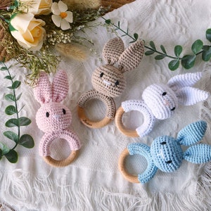 May include: Four crocheted bunny rattle toys with wooden rings. The bunnies are pink, brown, white, and blue.