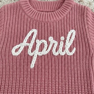 May include: A close-up of a pink knitted sweater with the word "April" embroidered in white. The sweater has a ribbed texture and a crew neck. The white embroidery is in a cursive font.